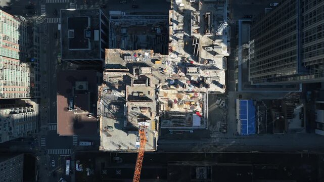 Top down showing crane building new skyscraper in downtown area, surrounded by tall office buildings with shadows and moving traffic on city streets below