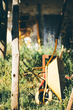 Selective focus outdoor detail of a yellow wheelbarrow and wooden post in grass, suggesting rural work, gardening, construction, and handmade countryside living
