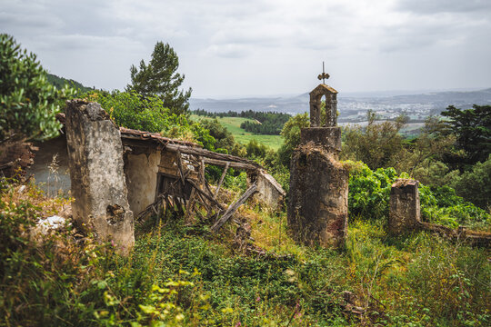 Weathered bell tower with cross stands beside shrubs and ruin walls, looking toward a wide countryside valley under a soft cloudy sky