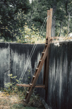 Close detail of a hand built wooden ladder and cable hardware inside an aged concrete pool, with posts, rustic surfaces, and soft forest background
