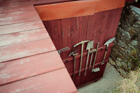 High angle rustic detail of red wooden boards, stone wall, blurred walkway, and hanging garden tools, ideal for rural workshop, storage, and texture themes