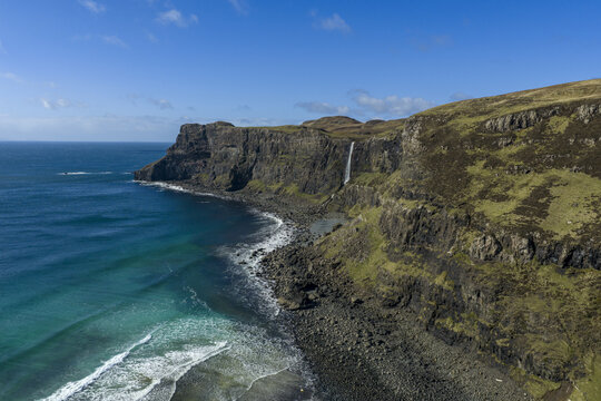 Aerial view of Talisker Bay Beach with its dramatic cliffs and a waterfall plunging into the Atlantic Ocean under a clear blue sky Isle of Skye, Scotland, United Kingdom.