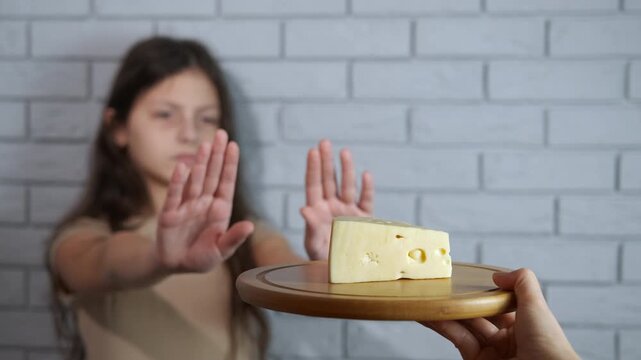 Dairy intolerance, avoidance of cheese. Unhappy young girl gesturing no with her hands and refusing a piece of cheese on a wooden board, illustrating the concept of lactose intolerance, dairy allergy