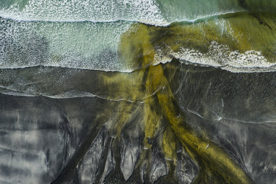 Aerial view of Talisker Bay Beach featuring dark volcanic sand and yellow river water mixing with crashing ocean waves Isle of Skye, Scotland, United Kingdom.