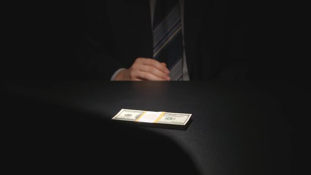 Anonymous businessman in a dark suit placing a thick stack of US dollar bills onto a black table under a dramatic spotlight, symbolizing corruption or a deal.