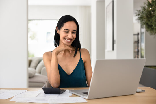 Smiling woman sitting at desk with laptop, calculator and papers, calculating expenses and organizing bills, feels satisfied after reviewing her budget, enough money and good savings for future plans