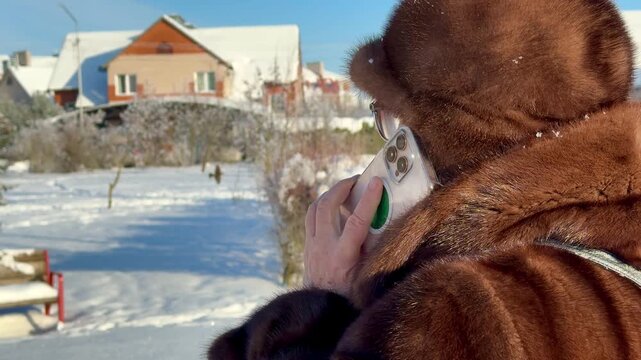 An elegant woman in a mink coat talks on her smartphone in a snowy park. A close-up of snow-dusted fur and a transparent phone case is visible on the side
