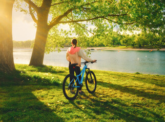 Obraz premium Woman riding a mountain bike near green trees and lake at sunset in spring. Colorful landscape with sporty girl, bicycle, beach, river, green grass, river in park. Summer. Sport and travel. Biking