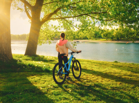 Woman riding a mountain bike near green trees and lake at sunset in spring. Colorful landscape with sporty girl, bicycle, beach, river, green grass, river in park. Summer. Sport and travel. Biking