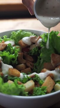 Fresh salad greens with crunchy croutons and chicken pieces. Hand pours creamy dressing onto the bowl. The dressing drizzles artfully over the ingredients