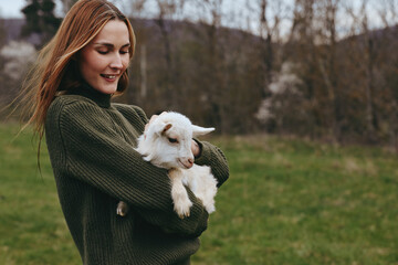 Obraz premium A woman in a dark green sweater holds a fluffy white goat kid in a green field, presenting a warm countryside lifestyle moment with gentle touch and peaceful animal companionship