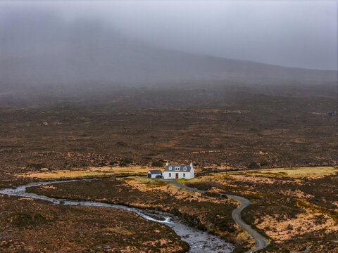 Aerial view of a white cottage nestled in the rugged moorland landscape with a winding road and stream under misty hills Isle of Skye, Scotland, United Kingdom.