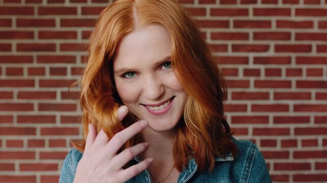 Happy girl, face and student playing with red hair for flirt or attraction on a wall background. Portrait, female person or fidgeting with curls or sign for romantic gesture, signal or expression