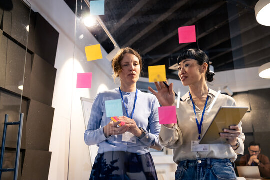 Diverse businesswomen collaborating efficiently, brainstorming ideas using sticky notes on glass wall and digital tablet in modern office.
