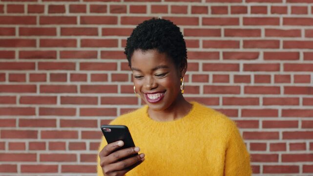 Happy, black woman and student with phone by brick wall for networking, social media or email for college. Smile, tech and person with cellphone for texting or feedback on university application.