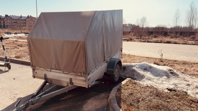 Gray tarpaulin trailer parked in lot, single axle utility trailer with weathered cover, visible hitch, wheel, rim and reflector