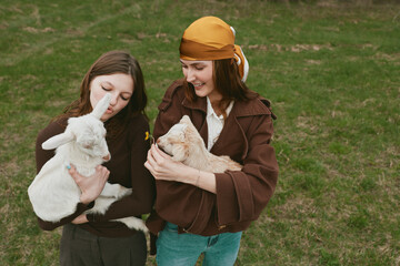 Obraz premium A pair of women share a gentle moment with a baby goat in an open grassy field, capturing a wholesome lifestyle scene focused on animal care, friendship, and outdoor farm life.