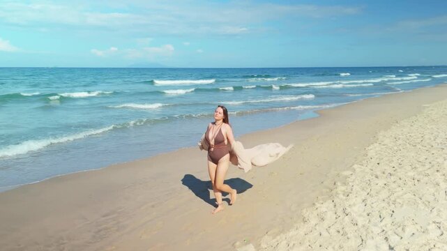 Woman walking barefoot on a golden sandy beach during a beautiful sunny day. Wearing a brown swimsuit and a beige sarong blowing in the wind. Experiencing serenity and freedom by the calm ocean waves