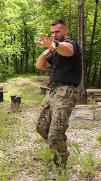 Man in camouflage tactical clothing making an overacted karate style hand move during outdoor martial arts practice, humorous combat themed footage filmed in a forest environment.
