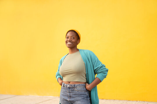 Smiling woman wearing knit cap and teal cardigan standing against bright yellow background, relaxed pose with confident expression in modern lifestyle scene