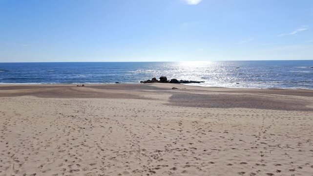 A wide angle view of Praia de Miramar in Portugal shows footprints on soft sand and rocky outcrops in the water. Bright sunlight creates a shimmering reflection on the calm blue Atlantic Ocean.