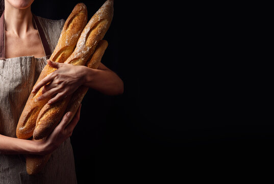 A person wearing an apron holds several freshly baked baguettes against a dark background.