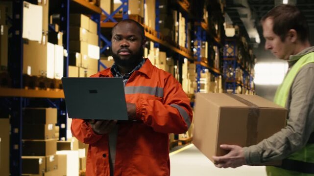 Diverse colleagues moving cargo in boxes using laptop for inventory control in large business warehouse. Distribution hub staff coordinate order fulfillment and shipping operations.