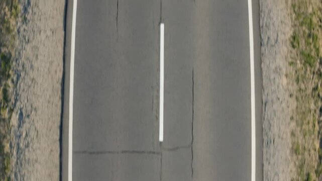 Aerial top-down view of an empty two-lane asphalt road with a central dashed white line, visible surface cracks, and gravel shoulders on both sides, filmed in daylight.