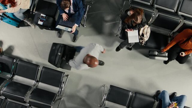 Passengers in busy airport create congestion carrying trolleys next to people sitting on chairs, top down view. Travelers in crowded air transportation hub walking with luggage and waiting on benches