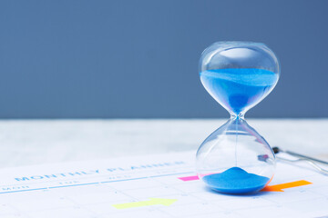Close-up of a monthly planner clipboard with highlighted dates using colorful arrows and a blue hourglass timer. Concept time management, scheduling, deadlines and productivity planning. © vadish