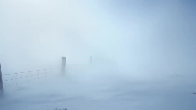 Intense snowstorm with heavy wind blowing snow across a desolate winter landscape, a rustic fence disappearing into a whiteout blizzard, representing extreme weather, isolation, and survival.