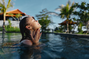 Fototapeta premium Young woman with wet hair relaxes in a sunlit pool, her face tilted toward the light as water ripples softly around her, a moment of tranquil summer bliss.
