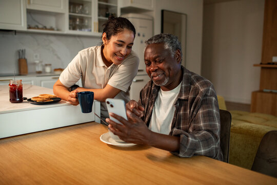 Caregiver helping senior man with smartphone at kitchen table