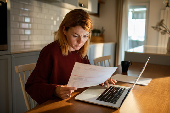 Focused woman reviewing bills on laptop at kitchen table