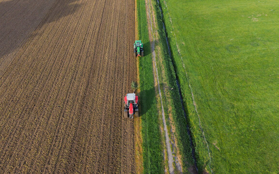 Aerial view of two tractors working on agricultural fields with plowed soil and green grass under bright sunlight Vierpolders, South Holland, Netherlands.