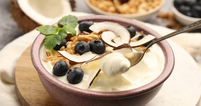 Eating tasty yogurt with coconut, blueberries, nuts, oatmeal and melissa at table, closeup