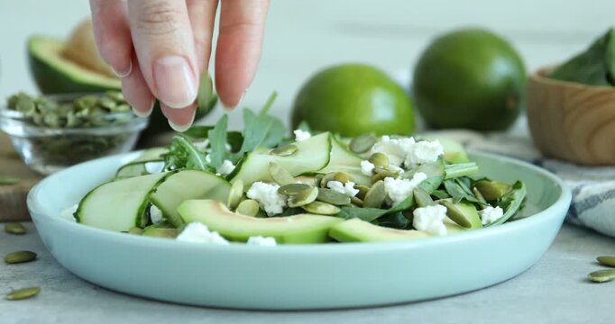 Woman adding pumpkin seeds to green salad with cucumber, avocado and feta cheese at grey table, closeup