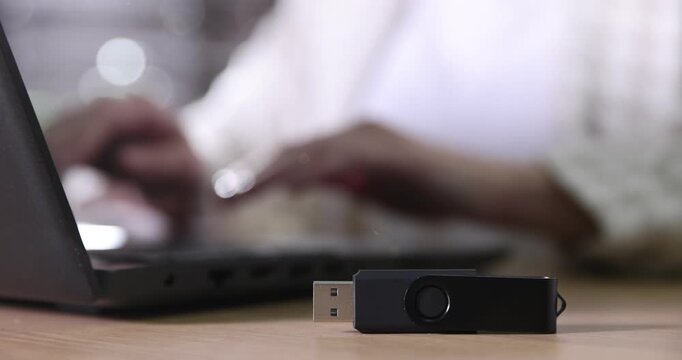 Woman using laptop at wooden table indoors, focus on flash drive