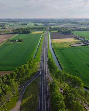 Aerial view of a straight highway with cars passing through vibrant green fields, lined with tall trees and a parallel canal under a clear sky.