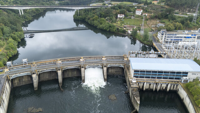 Aerial view of Subestacion Electrica Velle and the Velle Dam with water rushing through the spillway on the Minho River surrounded by green hills Ourense, Ourense, Spain.