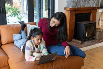 Naklejka premium Indian mother and daughter sitting on leather sectional sofa at home sharing tablet and headphones