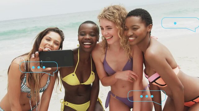 Four women extending phone, posing on shore adjusting for social selfie while chat bubbles floating