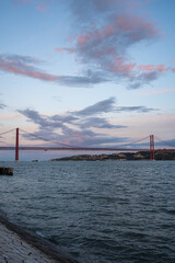 Obraz premium Suspension Bridge Over Wide River at Sunset with Dynamic Clouds and Calm Water Views
