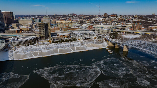 Aerial view of the snow-covered riverfront with ice floes on the water and the city skyline under a clear blue sky in Cincinnati, Ohio, United States.