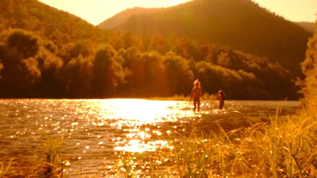Blurred sunset nature, soft blurred silhouette of a teenage girls walking and splashing water, standing in shallow water in mountain river at sunset. Refraction reflection sunlight. Cinematic effect