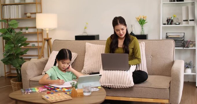 Mother working on laptop at coffee table while daughter focuses on drawing and learning nearby, illustrating remote work, distance education and family bonding in a cozy living room