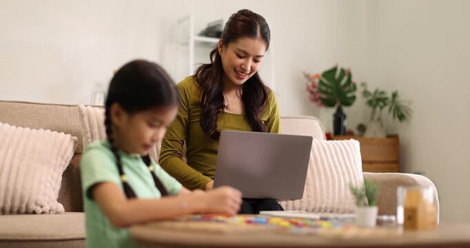 Asian mother sitting on a sofa and focusing on a laptop, working from home while her young daughter is playing with a puzzle game on a low table in the living room, depicting work-life balance
