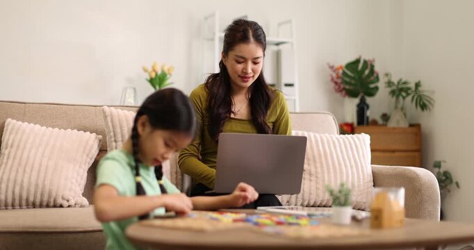 Asian mother sitting on a sofa and focusing on a laptop, working from home while her young daughter is playing with a puzzle game on a low table in the living room, depicting work-life balance