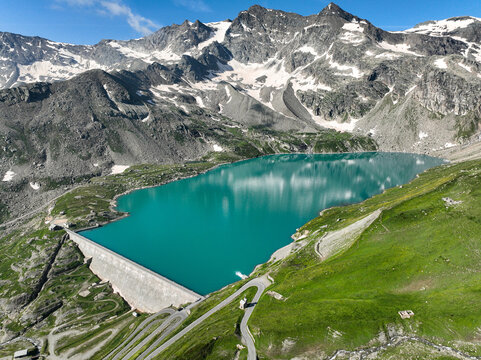 Aerial view of Serru Lake with its turquoise water, massive concrete dam, winding mountain roads, and snow-dusted peaks under a clear sky Ceresole Reale, Piedmont, Italy.