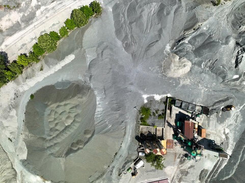 Aerial view of large grey gravel piles and industrial processing machinery at a quarry site with green trees along the perimeter in Caprie, Piedmont, Italy.